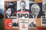 Wahlplakate für die Bundestagswahl 1965 kleben auf einer Holzwand an einer Straße.