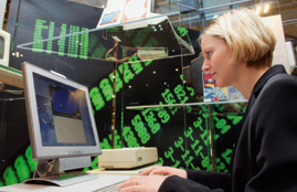 Frau vor Computerbildschirm, dahinter an der Wand Zahlenkolonnen