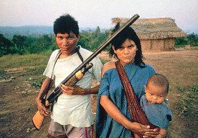 südamerikanisches Paar vor einer Hütte, die Frau hält ein Baby auf dem Arm, der Mann ein Gewehr