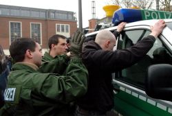 2 Polizisten überprüfen einen Rechtsextremisten, der gegen ein Polizeiauto gedrückt wird.