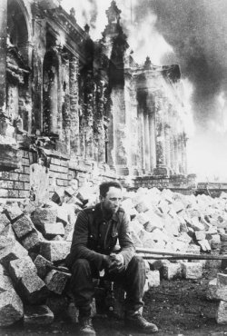 Fotografie: Deutscher Soldat auf Trümmern sitzend vor zerstörtem Reichstagsgebäude.