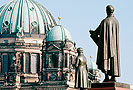 Bild: Kuppel des Reichstagsgebäudes. Davor zwei Menschen auf der Brücke zwischen Marie-Elisabeth-Lüders-Haus und Paul-Löbe-Haus.