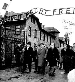 Fotografie: Die Gruppe des Frankfurter Schwurgerichts und zahlreiche Journalisten passieren das Lagertor mit der Aufschrift "Arbeit macht frei".