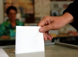 Foto: Eine Hand, die einen Stimmzettel hält, der bereits im Schlitz einer Wahlurne steckt. Im Hintergrund, unscharf, eine Person