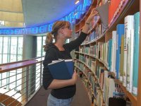 Foto: Benutzerin der Bibliothek hält mehrere Bücher in der Hand und nimmt ein weiteres Buch aus dem Regal