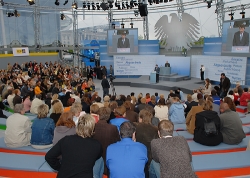 Besucher sitzen in der Bundestagsarena und hören einem Redner zu.