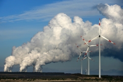 Windkrafträder stehen vor einem Kohlekraftwerk mit rauchenden Schornsteinen