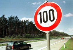 Foto: Verkehrszeichen, zugelassene Höchstgeschwindigkeit ist 100km/h, im Hintergrund eine Autobahn mit einem Auto und Wald