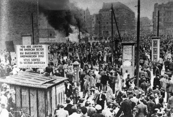 Menschen auf dem Potsdamer Platz, im Vordergrund ein Schild , das in drei Sprachen auf die Grenze des amerikanischen Sektors hinweist, im Hintergrund ein Feuer