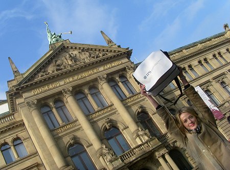 Eine junge Frau hält eine Tasche mit dem IPS-Logo hoch vor dem Museum Alexander König.