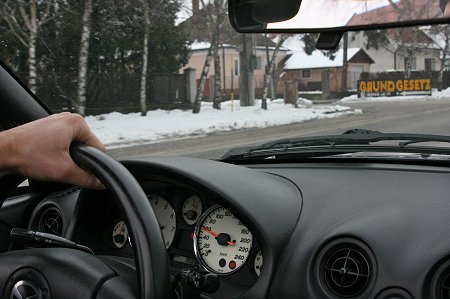 Hinter dem Lenkrad zeigt die Tachoanzeige eines Autos 60km/h an. Am Straßenrand ein Schild "Grundgesetz".