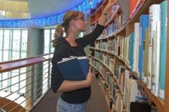 Foto: Benutzerin der Bibliothek hält mehrere Bücher in der Hand und nimmt ein weiteres Buch aus dem Regal