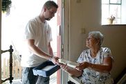 Ein Mitarbeiter des Pflegedienstes übergibt die verpackte Mahlzeit einer alten gehbehinderten Dame an der Wohnungstür