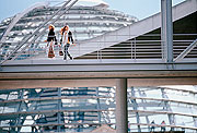 Bild: Kuppel des Reichstagsgebäudes. Davor zwei Menschen auf der Brücke zwischen Marie-Elisabeth-Lüders-Haus und Paul-Löbe-Haus.