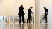 Bilder lehnen in der Ausstellungshalle des Kunst-Raums nebeneinander an der Wand, davor drei Personen