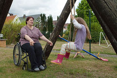 Eine im Rollstuhl sitzende Mutter spielt mit ihrer Tochter, die auf einer Schaukel sitzt. 