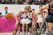 Sportlerin fährt mit ihrem Rollstuhl Rad an Rad mit einer Gegnerin dem Basketball hinterher.