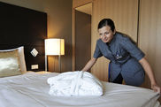 Ein Zimmermädchen richtet das Bett in einem Hotelzimmer. Auf dem Bett liegt gefaltet ein weißer Bademantel. 
