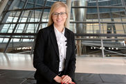 Eine junge Frau mit blonden, offenen Haaren, einem dunklen Sakko und einer weißen Rüschenbluse lächelt in die Kamera. Im Hintergrund ist sieht man in das Innere der Reichstagskuppel.