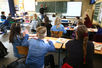 Blick von hinten in ein Klassenzimmer. Die Rücken der Schüler im Vordergrund, ein Lehrer an der Tafel im Hintergrund. 