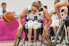 Sportlerin fährt mit ihrem Rollstuhl Rad an Rad mit einer Gegnerin dem Basketball hinterher.