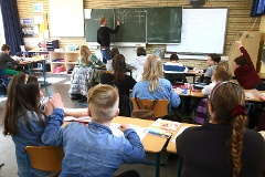 Blick von hinten in ein Klassenzimmer. Die Rücken der Schüler im Vordergrund, ein Lehrer an der Tafel im Hintergrund.