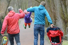 Ein Kind schaukelt an den Händen zweier Männer in der Luft. Mit der rechten Hand schiebt einer der Männer einen Kinderwagen.