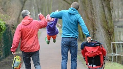 Ein Kind schaukelt an den Händen zweier Männer in der Luft. Mit der rechten Hand schiebt einer der Männer einen Kinderwagen.