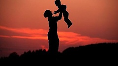 Eine Mutter hebt vor dem rot gefärbten Abendhimmel ihr Kind hoch