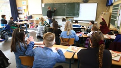 Blick von hinten in ein Klassenzimmer. Die Rücken der Schüler im Vordergrund, ein Lehrer an der Tafel im Hintergrund.