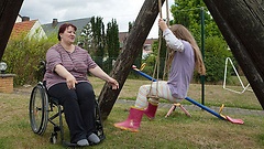 Eine im Rollstuhl sitzende Mutter spielt mit ihrer Tochter, die auf einer Schaukel sitzt.