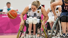 Sportlerin fährt mit ihrem Rollstuhl Rad an Rad mit einer Gegnerin dem Basketball hinterher.