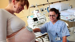 Eine Schwangere in einem Kreissaal. Am Bauch ist ein Wehenschreiber befestigt. Vor ihr steht eine Hebamme.